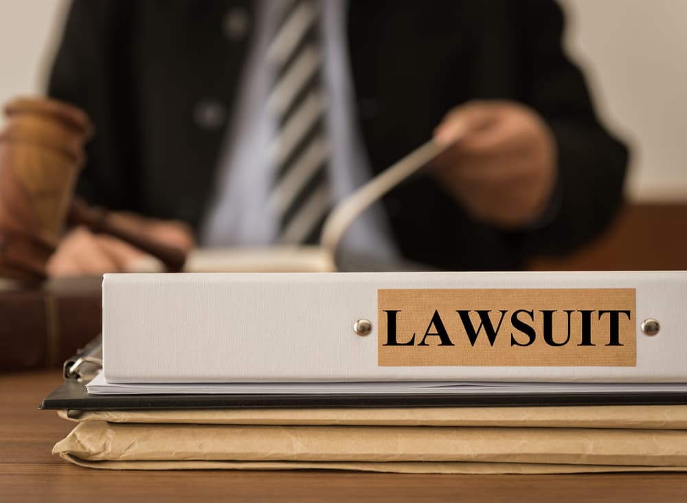Lawsuit file binder on desk with judge reviewing documents in the background.