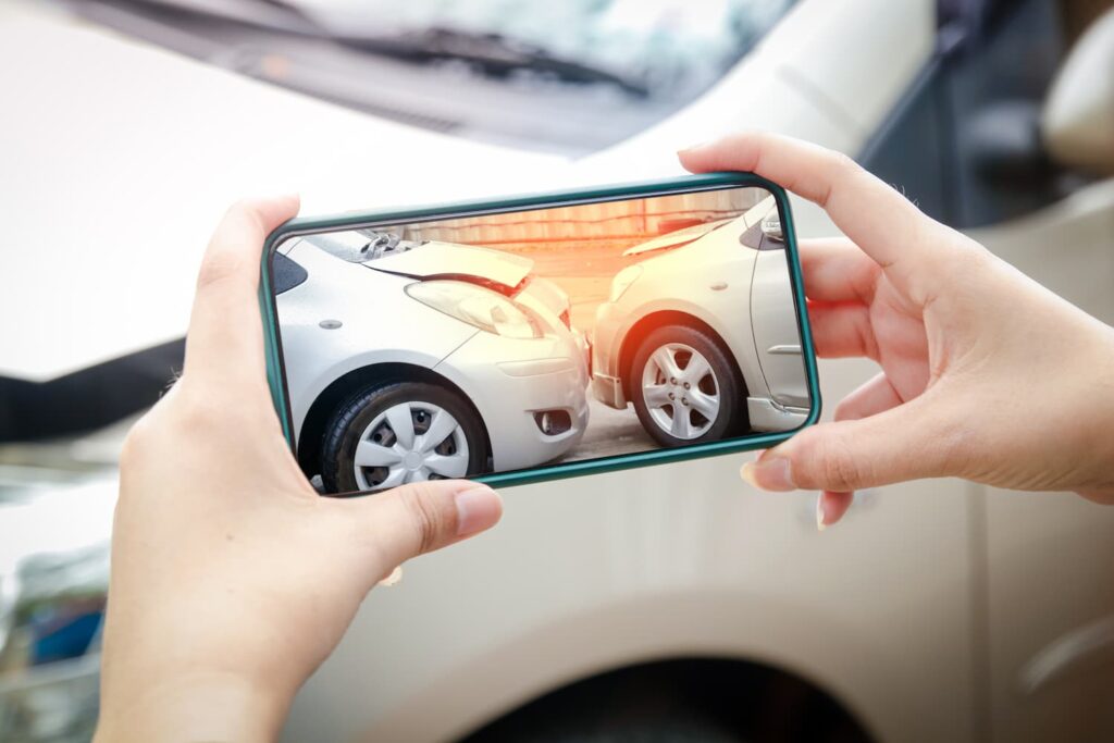 Driver using a smartphone to photograph vehicle damage after a car accident, documenting collision evidence for an insurance or injury claim.